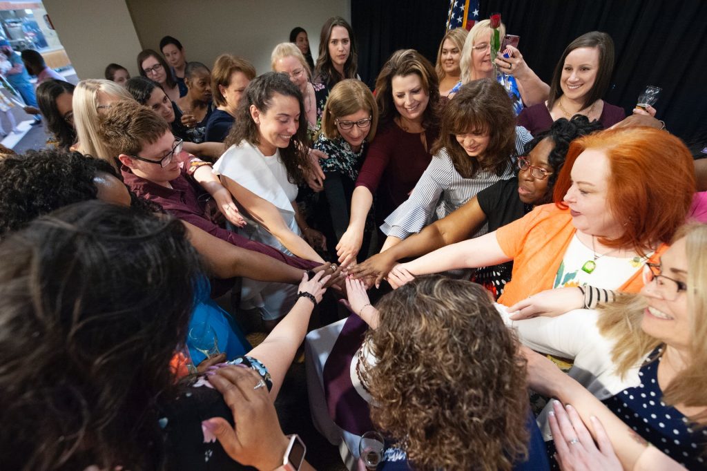 This is a color photo of a group of women standing in a circle with their hands together in the middle.