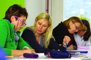 This is a color image of four adult women sitting at a table working. The women are working in pairs. There are notebooks, pens, and books on the table.