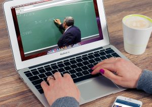 This image shows a set of hands hovering over a laptop keyboard. On the screen there is a man standing in front of a green chalkboard writing out an equation.