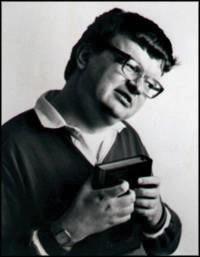 This is an image of Kim Peek, the subject of the movie Rain Man. He is shown in a black and white photo, holding a book up to this chest. He is wearing glasses and a long-sleeved polo shirt