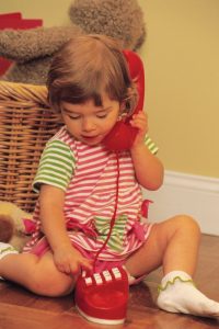 This image shows a female toddler sitting on the floor holding a pretend phone to her ear while dialing the numbers with her other hand. She is wearing a pink and green striped shirt with pink shorts and white socks. The pretend phone she is holding is red.