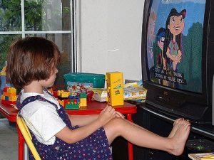 This is a photo of a young female child sitting in a chair with her feet up resting against a media center. She is watching a cartoon on television.