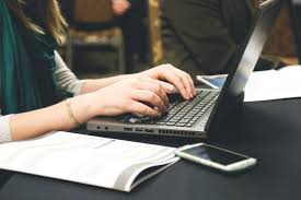 This is an image of a woman typing on a laptop. The individual is sitting at a desk with both hands on the keyboard. The laptop is black and silver. The person is wearing a green scarf and several gold bracelets on her right wrist. There is a cell phone and paper booklet to the right of her laptop. The image only shows the person's arms, hands and part of the torso.