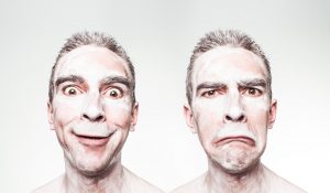 This image shows two identical figures standing side-by-side, each making a different facial expression. The figure is a male and the photo is taken from the shoulders up. The figure has short hair, and he is covered in a white powdery substance. In the image on the left the figure’s mouth is turned upwards in the shape of a smile, and his eyes are opened wide. The image on the right shows the figure with his mouth facing downwards, eyes squinted, and furrowed brow.