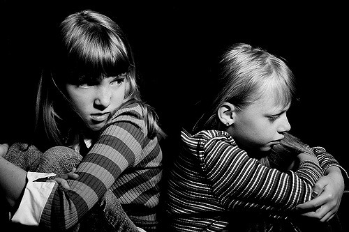 This is a black and white photo of two little girls sitting back to back. They are each wrapping their knees up with their outstretched arms. The girl on the right is slightly taller and she is looking over her left shoulder and making a scowling face in the direction of the other girl. She is wearing a striped shirt and pants. The girl on the right is wearing a striped shirt and she has a concerned look on her face and she is looking downwards and away from the other girl.