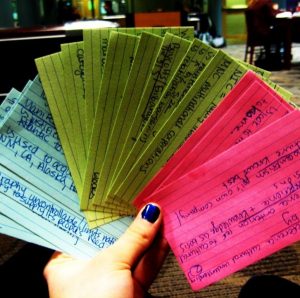 This image shows a left hand with painted blue nail polish holding a fanned stack of index cards. The cards on the left are blue; the cards in the middle are green; and the cards on the right are red. Each of the cards have handwriting on them in blue pen.