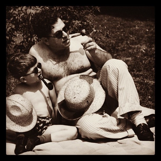 This image is in black and white and shows an adult male and a young, male child sitting outside on a blanket. Neither figure is wearing a shirt, and the young child is wearing shorts and the adult is wearing pants. Each figure is holding a tobacco pipe in their mouth, they each have a straw hat resting on their lap, and they are both wearing sunglasses.