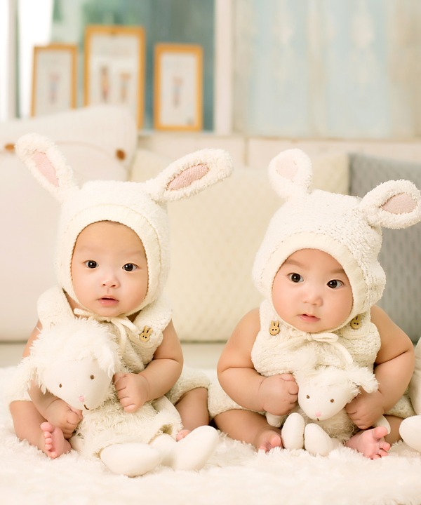 This image shows two identical twin babies sitting up side-by-side. They are both wearing a white bunny costume and holding a stuffed, white bunny toy. They are sitting on a fluffy white carpet and staring straight towards the camera.