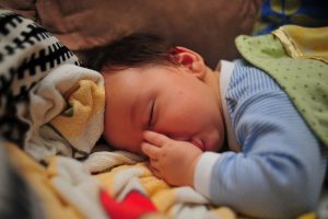 This is an image of a young infant lying face down in a pile of blankets. The baby is sleeping and has it’s left thumb in it’s mouth.