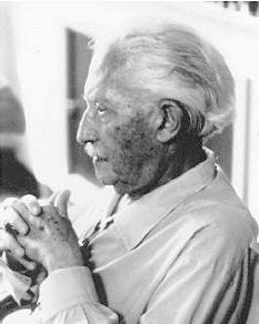 This is a black and white image of an elderly Erik Erikson. The image is a profile view of him sitting down with his elbows resting on the arm rest, hands folded together at his chest. He is looking forward.