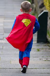 This image shows a young male child wearing a superman costume. He has a red cape on and he is walking away from the camera. He is on a stone walkway.