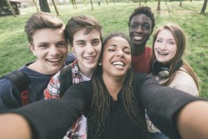 This is image shows five teenagers standing together taking a selfie. There are three males and two females. There are two, Caucasian males on the left, an African American woman holding the camera in the middle, an African American male standing in the back, and a Caucasian female standing on the right. They are standing outside surrounded by trees. All five of the teenagers are smiling.