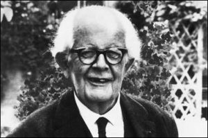 This is a black and white image of Jean Piaget. He is standing outside wearing a suit and tie. He is wearing glasses and smiling towards the camera.