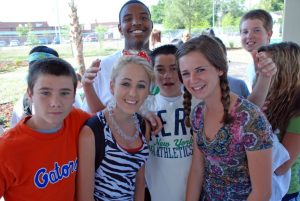 This is an image of seven teenagers standing outside. From left to right there is a male, a female, a male, and another female. Behind these individuals there are two more males and a female in the background. One of the females is wearing a backpack.