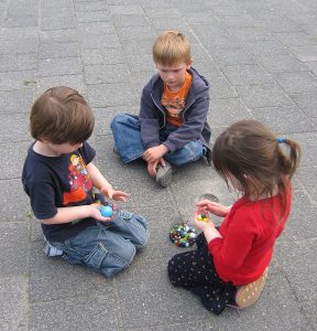 This image shows three young children sitting outside on the pavement. There is one little girl and two little boys. The little girl is holding marbles in her hand, and one of the little boys is also holding a larger blue ball along with a few marbles. The second little boy is watching.