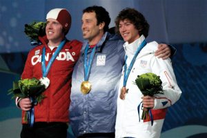 This image shows three male athletes standing side-by-side with their arms wrapped around one another while posing for a photo. The male on the left is wearing a red and white coat that says “Canada” across the front. He is wearing a red and white swimming cap, holding green flowers in his right hand, and wearing a silver medal around his neck. The male in the middle is wearing a blue and white coat with “USA” written on the upper left chest. He is wearing a gold medal around his neck. The male on the right is wearing a white coat that says “France” written on the upper left chest. He is holding green flowers in his left hand and wearing a bronze medal around his neck.