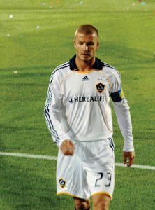 This image shows David Beckham walking across the soccer field. He is wearing a white uniform with blue and yellow accents. His jersey number is 23, which is also written on his shorts.