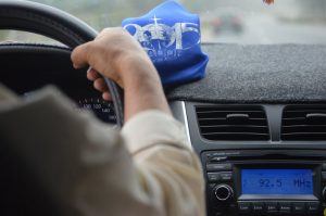 This image shows the right arm of a driver of a vehicle. The person’s right hand is holding onto the steering wheel as though they are driving. The radio is on and there is a blue cloth resting on the dashboard.