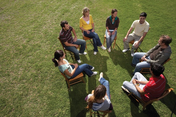 Group therapy provides a therapeutic setting where people meet with others to share problems or concerns, to better understand their own situation, and to learn from and with each other. This image shows a group of eight individuals, both male and female, sitting on chairs outside in the grass