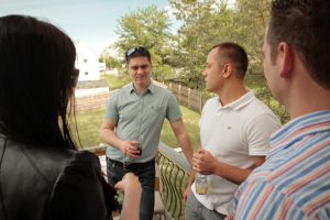 This image shows three adult men and one female standing outside on a deck socializing. There is a female figure in a black top facing away from the camera towards two other adult men. There is a third male figure in the image who is wearing a peach and blue striped shirt who also has his back towards the camera and is facing the other two men. The two men facing the camera are each holding a drink; the male on the right is holding a beer bottle and the male on the right is holding a clear glass with brown liquid. The male on the left is wearing a green, button-up shirt, jeans and a black belt. He is resting his left arm on the banister of the deck. The male on the right is wearing a short-sleeved, white polo shirt with gray pants.