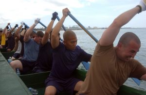 This image shows eleven men sitting in a crew boat in water. They all have their oars positioned above their head on their left side, with the oar facing downwards towards the water. Their positions are aligned so their rowing is synchronized.