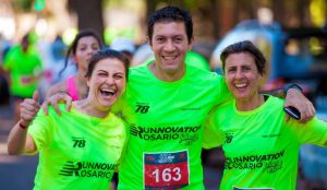This image shows three adults standing outside during a road race. There are two female figures on each end, and a male figure standing in between them. They have their arms wrapper around one another. The participants are wearing bright yellow event shirts with the racing bib pinned to the front. The three individuals are smiling, and the woman on the left is giving the “thumbs up” with her right hand.