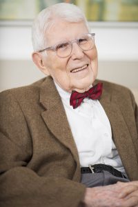 Aaron Beck and Albert Ellis were pioneers in cognitive therapy. The image is a color photo of Aaron beck. He is wearing a brown suit, with a collared white shirt, a red bow tie, and glasses. He is sitting down with his hands on his lap and smiling at the camera.