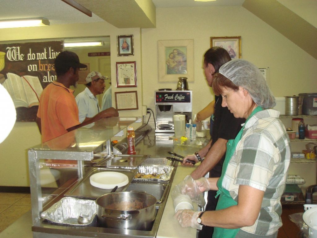 This image shows a male and female working behind the serving counter at a soup kitchen. The woman is wearing a hair net and gloves while taking cups out of the plastic wrapper. She is also wearing a checkered shirt with a green apron. There are two male figures standing by the counter waiting to be served.
