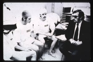 This is a black and white image of the Stanford prison experiment. In the image there is an adult male sitting on the right side of the room wearing a suit, tie and sunglasses. He has a pad of paper on the desk in front of him, and he is holding his hands by his waist. There are three adult males sitting in front of him, each wearing hat and white uniform. The three men have numbers written across their chest.