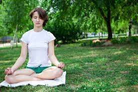 This image shows a young woman sitting on a blanket, outside in the grass. She has her legs crossed and her hands resting, palm-side up, in the meditative position. Her eyes are closed. She is wearing a white shirt and green shorts.
