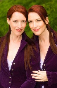 This image shows a picture of two, adult female identical twins. They are standing side by side, pictured outside with a green background. The image is taken from their waist up. The women have long, red hair pulled back halfway. They are wearing a purple, button-up coat with a lilac shirt underneath. The twin on the right side is resting her left hand on her right forearm.