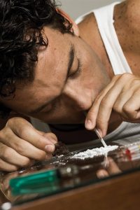 This image shows an adult male snorting cocaine off of a table. He is wearing a white tank top and he is leaning over the table, with his nose positioned over a straw and line of cocaine. There is a green lighter on the table.