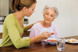 This image shows a young woman feeding an elderly woman. They are sitting at a table with a bowl of tomato soup, a glass of water, and a plate with some bread on it. The young woman is sitting on the left, wearing a green sweater. She has light brown hair that is pulled up into a pony tail. She is resting her left hand on the back of the elderly woman while holding a spoonful of soup. The elderly woman is wearing a pink shirt with a white napkin tucked into the collar. She has short gray hair and is wearing earrings.