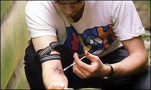This image shows an adult male squatting down while injecting his right arm with heroin. He is wearing a white, printed tee shirt and dark pants. He has his belt tied around his right bicep, and he is holding a syringe with his left hand. His forearm is bruised at the site of injection.