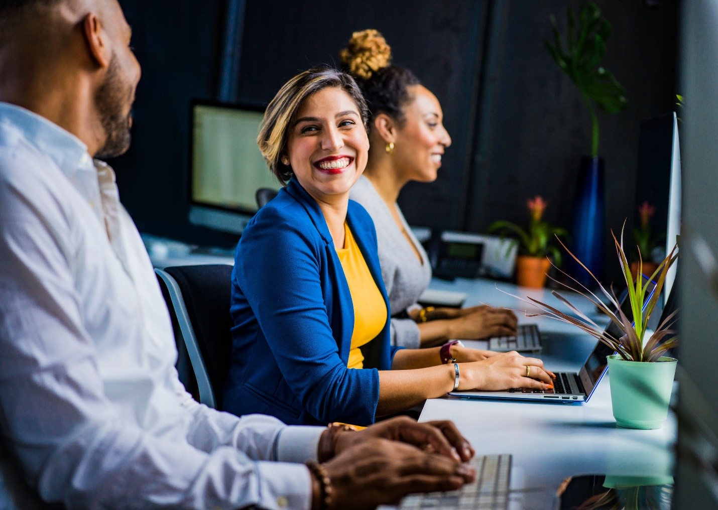 This is an image of three adult business professionals, sitting at a counter with laptops open. The woman in the middle is looking toward the camera and smiling at a co-worker. She is wearing a bright blue jacket with a vivid yellow shirt.