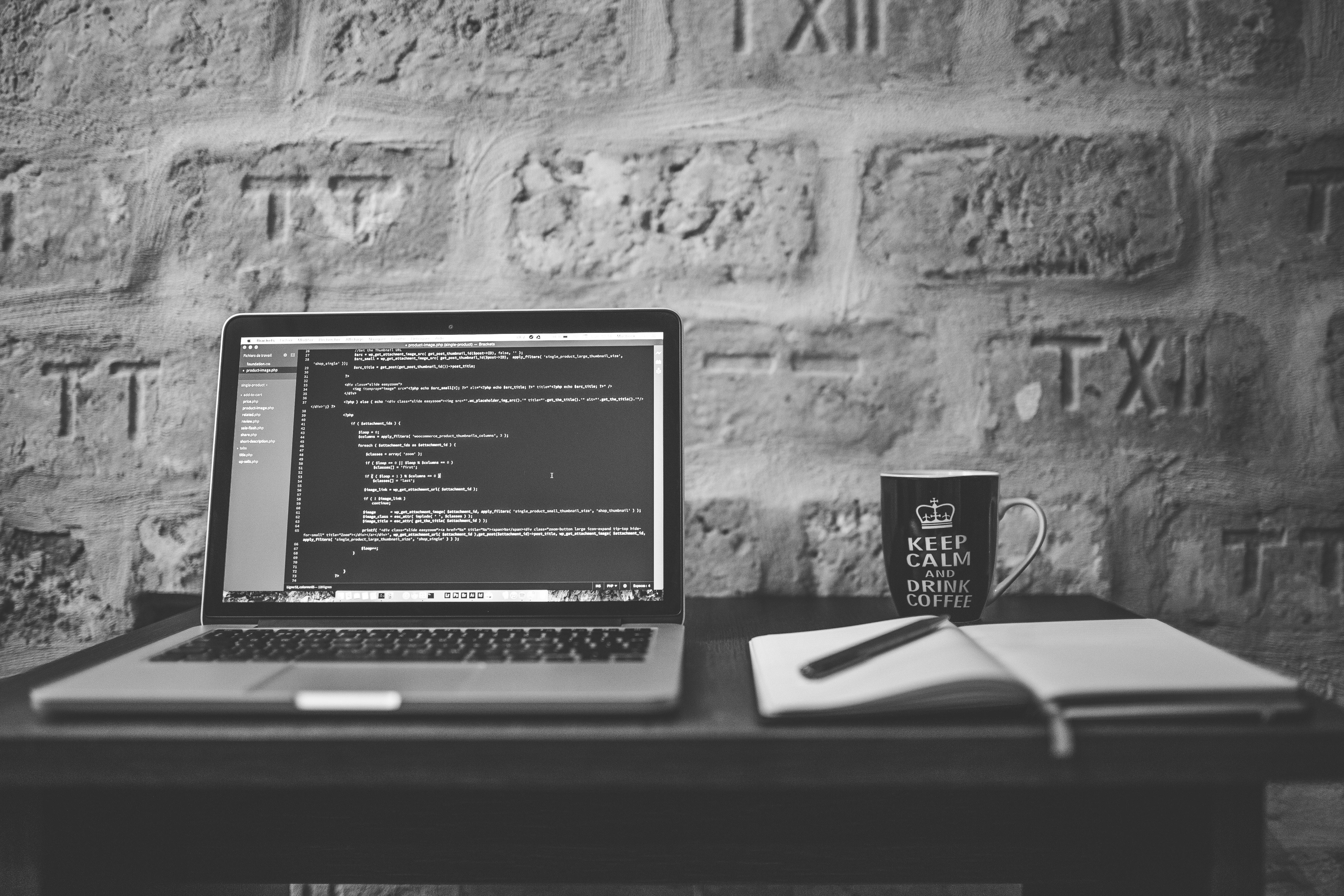 This is a gray-scale image of an open laptop on a wooden desk. The laptop has CSS code on the screen. There is a coffee mug next to the laptop that reads, keep calm and drink coffee.