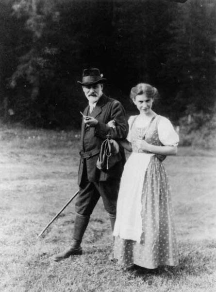This is a black and white photograph of Sigmund Freud and his daughter, Anna, walking outside in the grass. Sigmund is on the left and Anna is on the right. Sigmund is carrying a walking stick in his right hand and a pipe in his left hand. Anna is holding her father's left elbow with her right hand. She is wearing an ankle length dress with an apron.