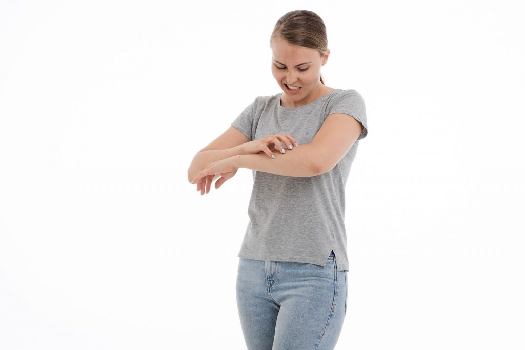 This is a color image of a woman scratching her left forearm with her right hand. She is wearing a gray t-shirt and jeans, and she is looking down at the arm she is scratching.