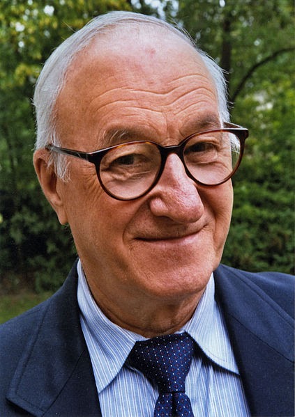 This is a color image of Albert Bandura. The image shows him standing outside as he poses for the camera. He is wearing a blue collared shirt, a tie and a suit coat.