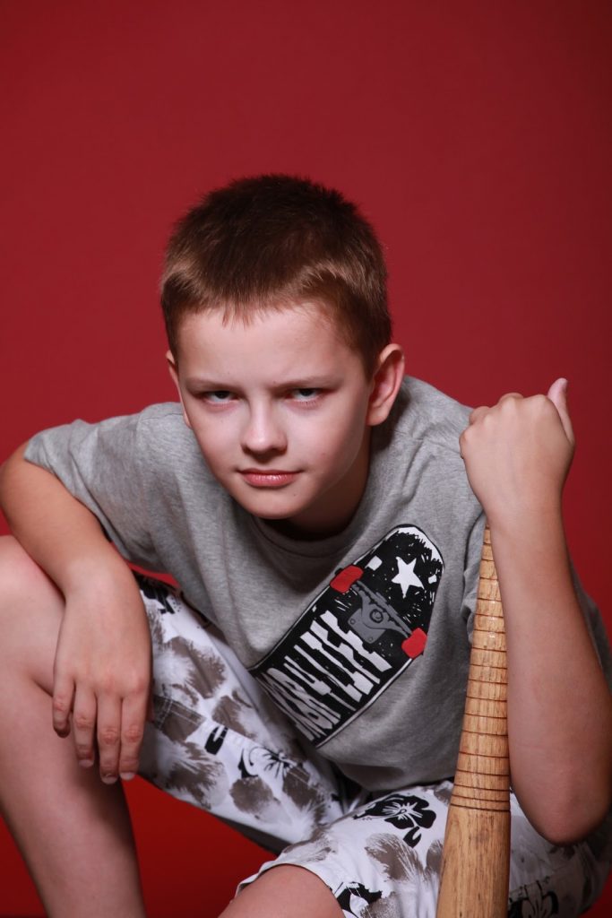 This is a color image of a boy kneeling on the ground while resting his left arm on a baseball bat.