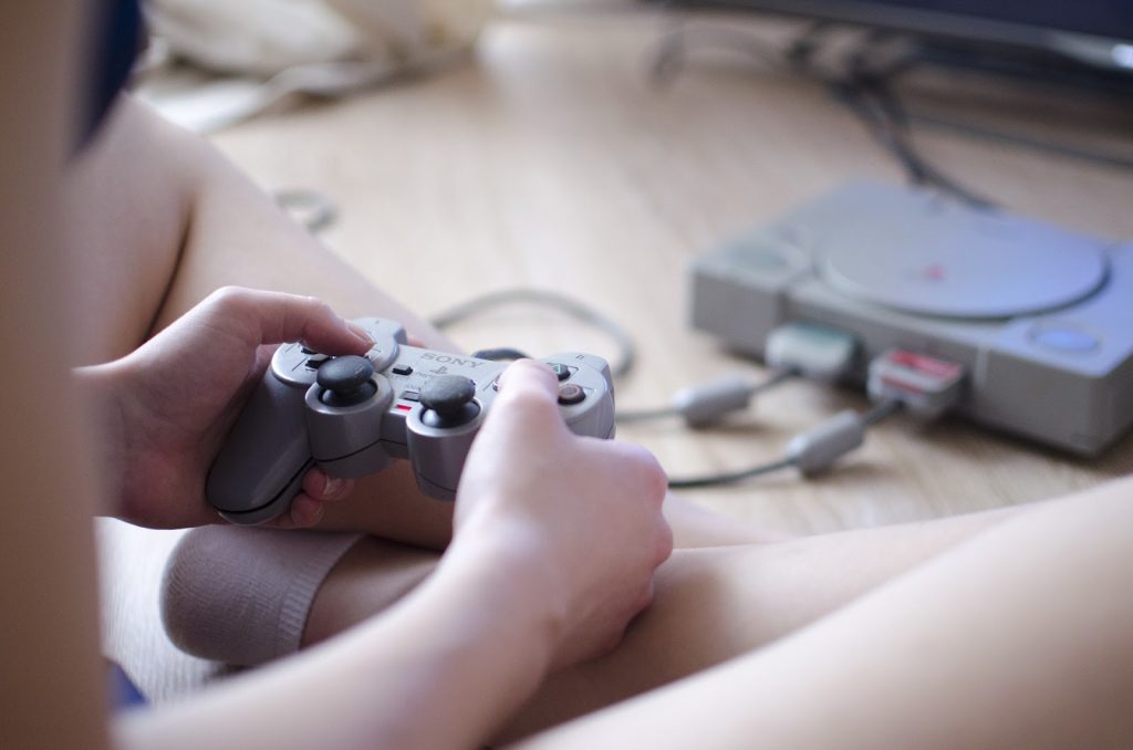 This is a color image of a child sitting on the floor playing a video game. The photo shows the child's legs and arms only as he/she holds a game controller in their hands. A video game console sits on the floor in front of the child.