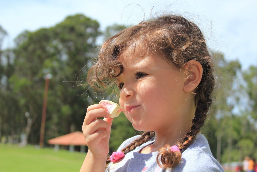 This image shows a little girl standing outside eating a marshmallow. She has her hair in braided pigtails and she is wearing a purple shirt.