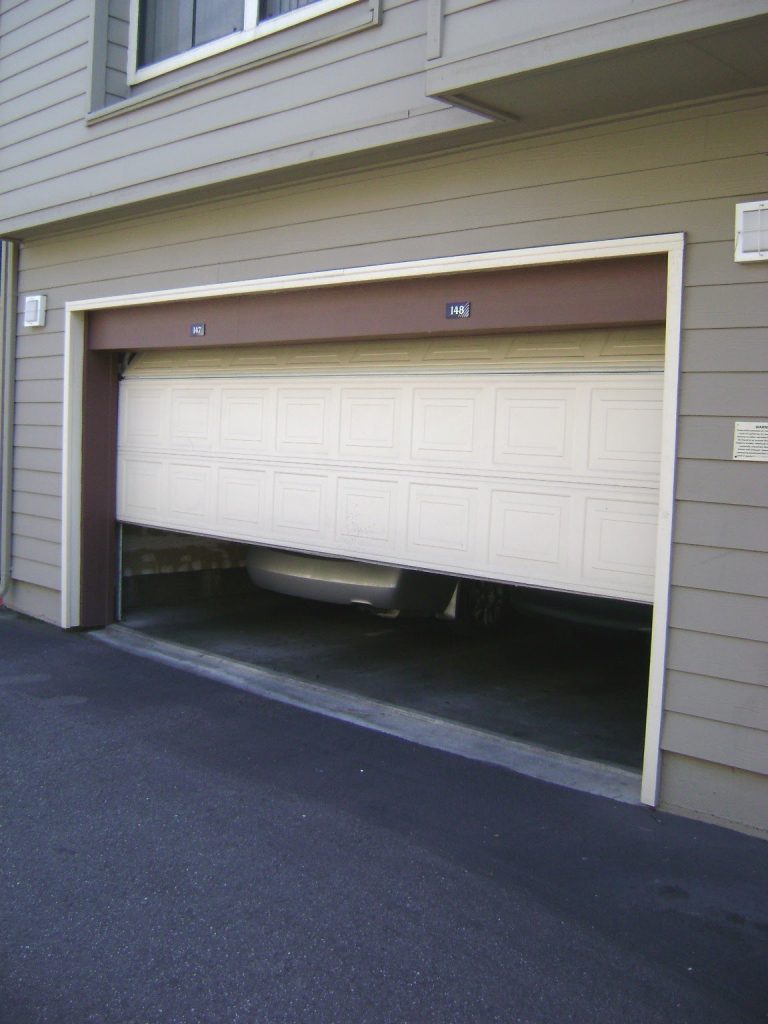 This is a color photograph of a garage door. The door is partially open and stuck. The bumper of the car parked in the garage is slightly visible.