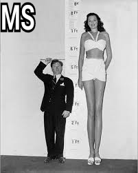 This is a color photograph of a tall woman and a short man. They are standing side-by-side in front of a wall-mounted measuring stick. The woman is wearing a white crop top and white shorts, and the man is wearing a black suit and tie. The woman is just under 8 feet tall, and the man is just over five feet tall.