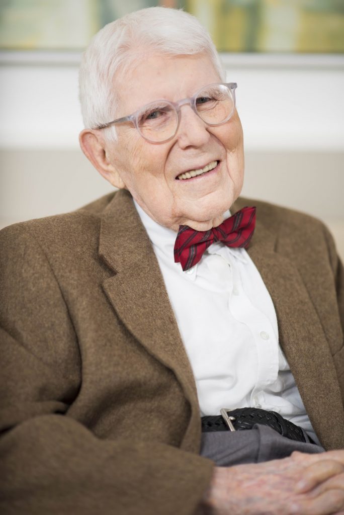 This is a color photograph of Aaron Beck. He is sitting down as he poses for the camera. He is wearing a brown suit coat, white collared shirt, gray pants, and a red bow tie. His hands are folded in his lap as he smiles for the camera.