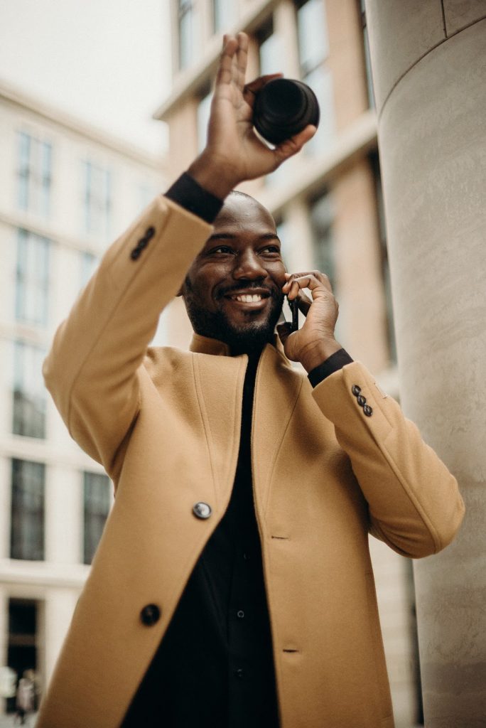 This is a color photograph of a man standing outside while talking on his cell phone. He is waving to someone with his right hand, as he holds his phone up to his ear with his left hand. He is wearing a tan jacket and a black top.