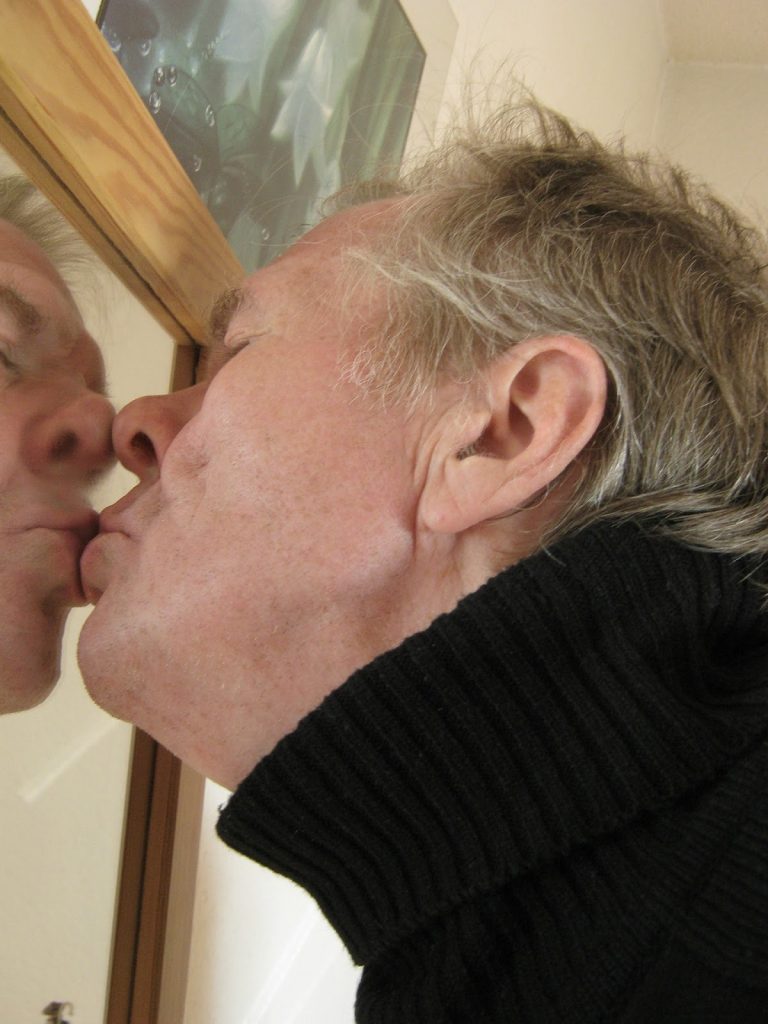 This is a color image of a man kissing his reflection in a mirror. He is wearing a black turtle neck top.