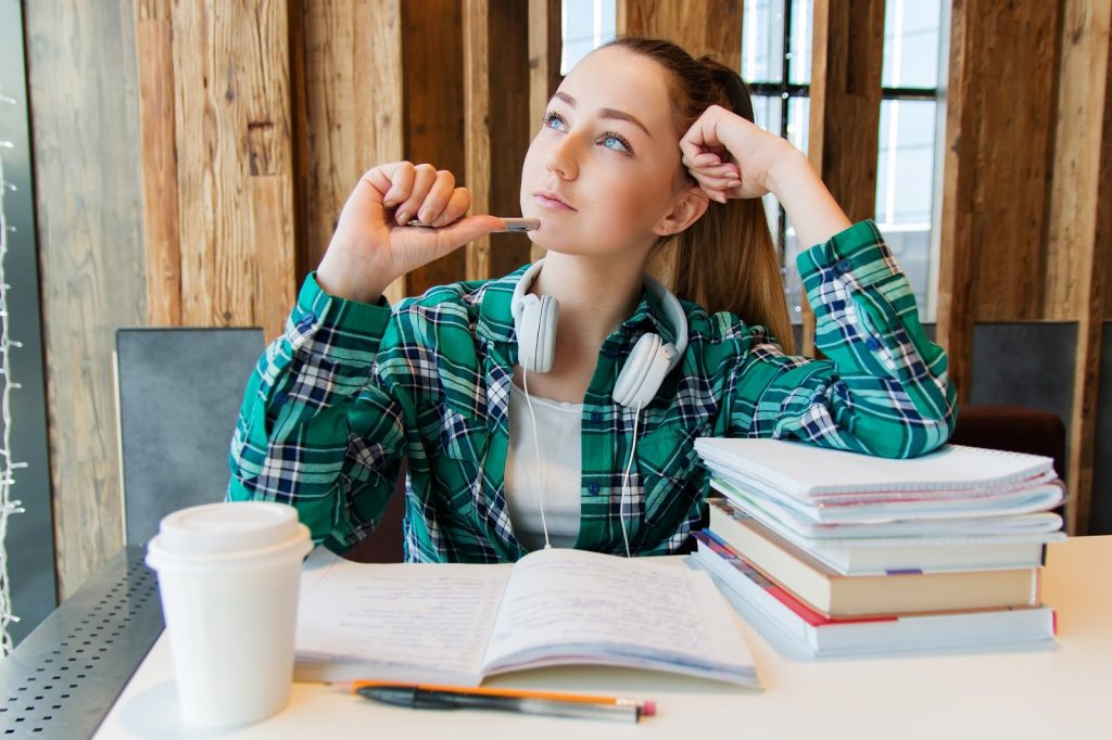 This is a color image of an adolescent girl sitting at desk completing her homework. She has a stack of books in front of her, as well as a coffee cup and a few pens and pencils. She is holding a pencil up to her chin as she looks up while pondering.