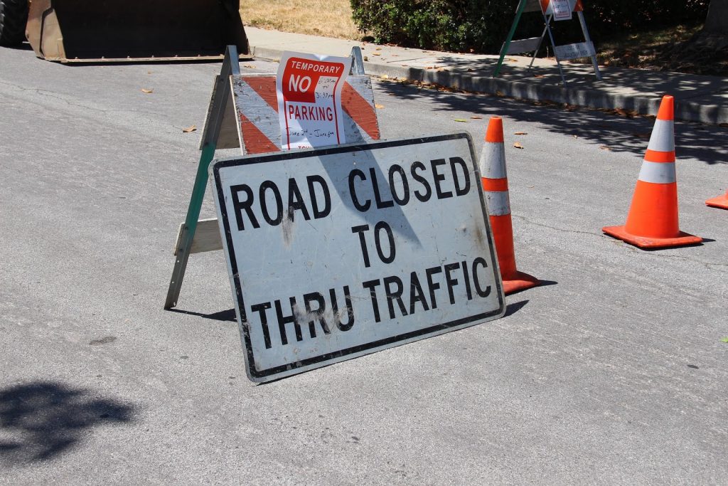 This is a color image of a detour. There are two yellow traffic cones and a white sign that reads "Road Closed to Thru Traffic."