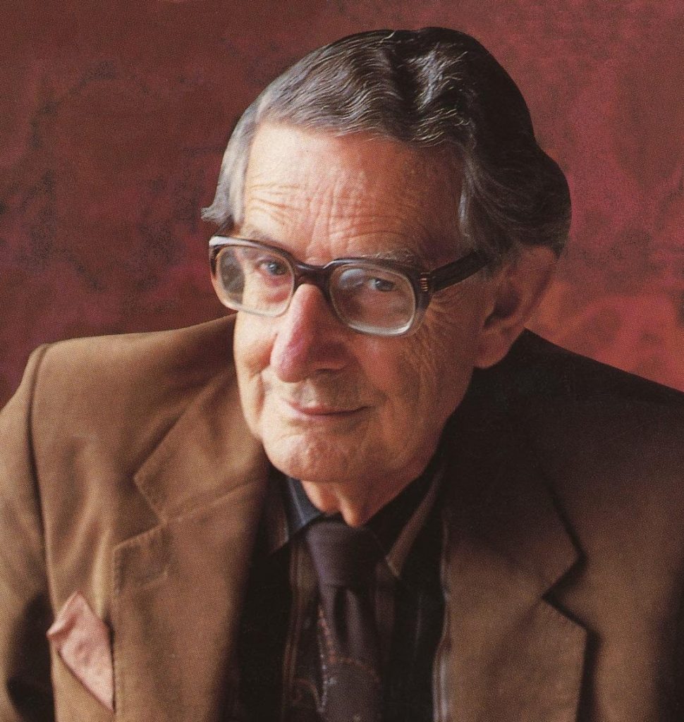 This is a color photograph of Hans Eysenck. He is smiling for the photo while wearing a brown suit coat, brown shirt and tie, and glasses.